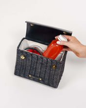 Open view of the Black Wicker Mini by Modern Picnic, showing the insulated interior filled with a container of fresh strawberries and other food items, while a hand places a red juice bottle inside, against a clean white background.