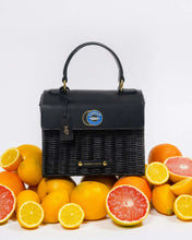 Black Wicker Caviar Mini by Modern Picnic styled in a citrus fruit display, featuring whole and sliced lemons, oranges, and grapefruits arranged around the insulated wicker bag with a black vegan leather flap, gold hardware, and blue caviar emblem on a clean white background.