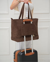 Woman showing the smart luggage sleeve feature of a brown suede tote bag, securely slid over the handle of a black rolling suitcase.