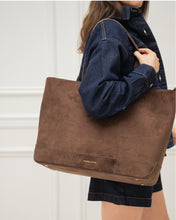 Woman carrying a large brown suede tote bag on her shoulder while wearing a dark wash denim jacket for a casual fall fashion look with a blurred white background.