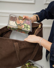 Person placing a container of salad into a brown suede tote bag with another container of salad on a white surface.