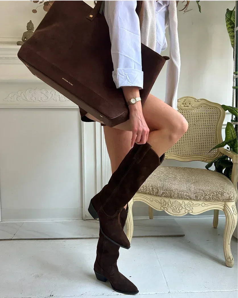 Stylish outfit featuring a woman holding a large brown suede tote bag and wearing matching dark brown suede knee-high boots and a white shirt indoors.