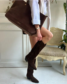Stylish outfit featuring a woman holding a large brown suede tote bag and wearing matching dark brown suede knee-high boots and a white shirt indoors.