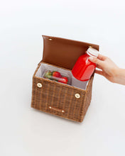 Open Brown Wicker Mini Lunchbox by Modern Picnic showing its insulated interior, with a hand placing a red juice bottle inside next to fresh strawberries.