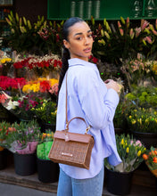 Woman wearing a light blue shirt and jeans with the Brown Wicker Mini Lunchbox by Modern Picnic styled as a crossbody while shopping at an outdoor flower market.