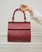 A hand holds the structured red smooth luncher by its curved top handle, showcasing the flap closure and horizontal gold-toned bar detail against a subtly textured light background.