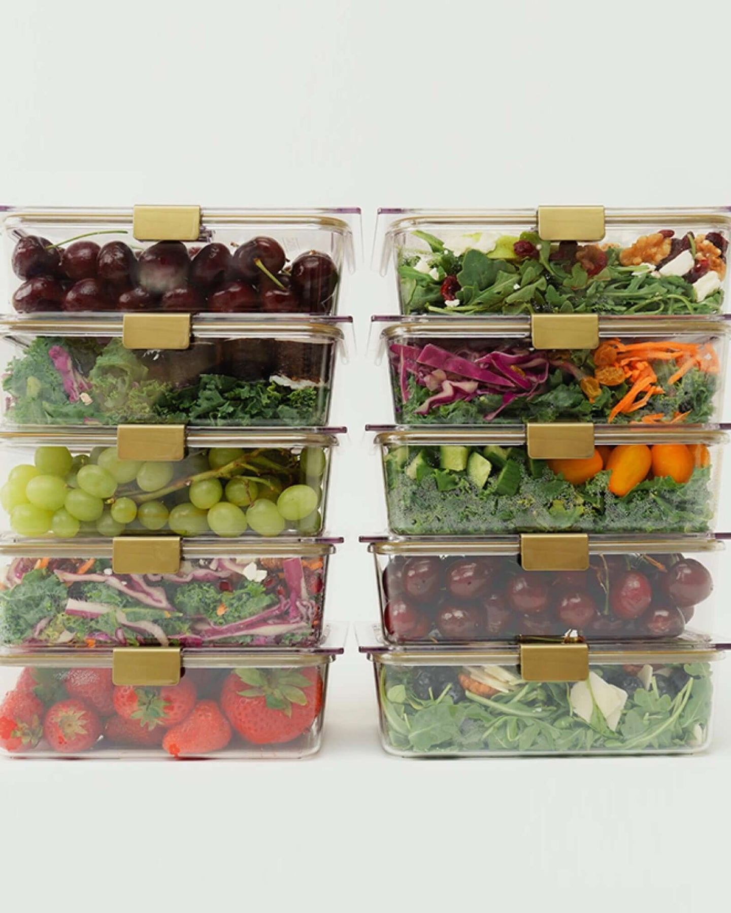 A studio shot showing ten clear, rectangular food containers stacked into two columns of five. The containers are filled with assorted salads (greens, carrots, cabbage) and fresh fruit (cherries, grapes, strawberries).