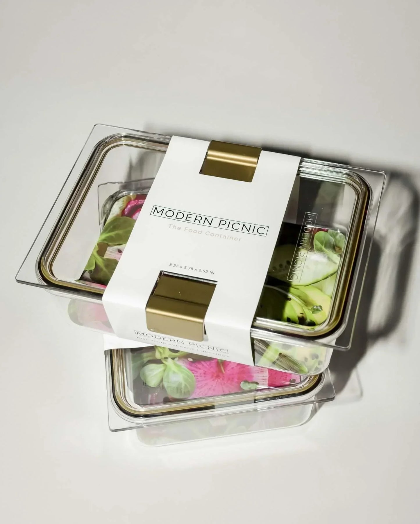 An angled studio shot of two clear, rectangular food containers stacked, wrapped with a white paper sleeve labeled "MODERN PICNIC The Food Container." The visible food inside includes greens and pink cabbage.
