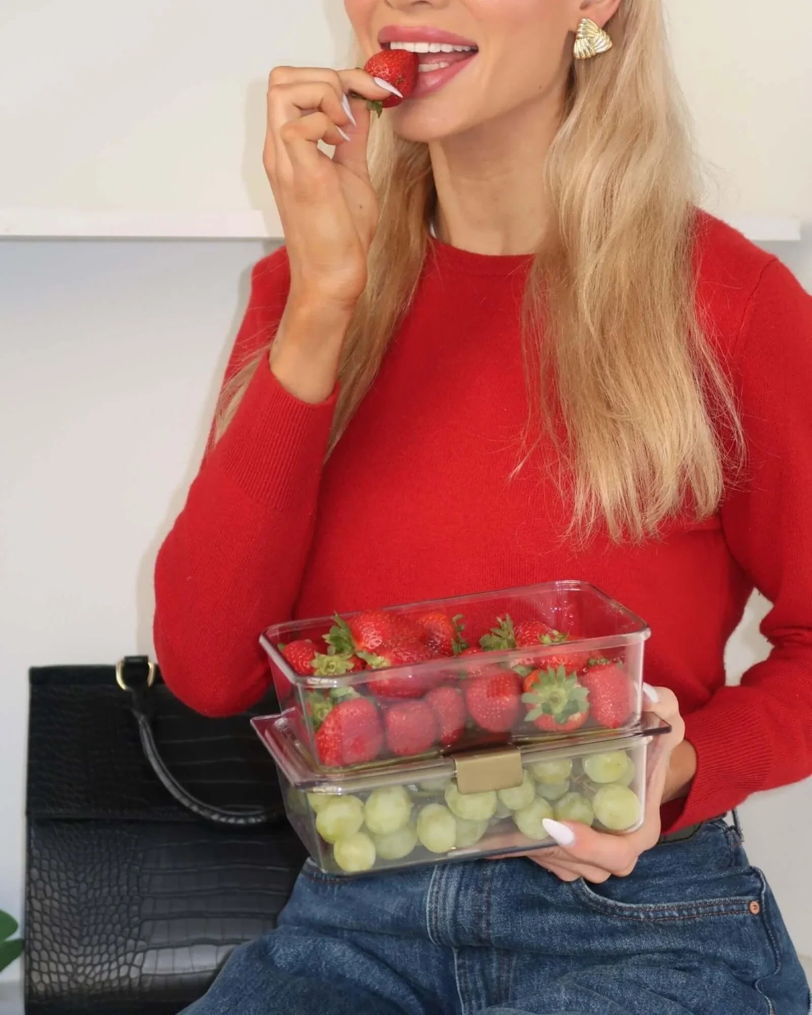 A woman with blonde hair, wearing a red sweater and blue jeans, is biting a strawberry while holding a stack of two clear containers. The top container is filled with strawberries, and the bottom container is filled with green grapes. A black handbag is visible in the background.