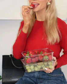 A woman with blonde hair, wearing a red sweater and blue jeans, is biting a strawberry while holding a stack of two clear containers. The top container is filled with strawberries, and the bottom container is filled with green grapes. A black handbag is visible in the background.
