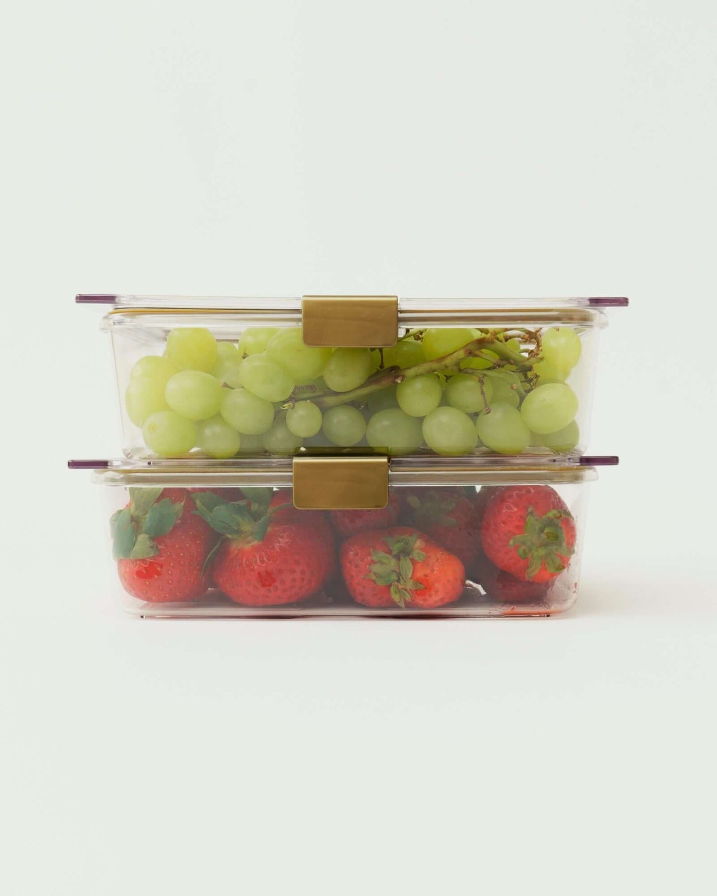 A studio shot showing two clear, rectangular food containers stacked vertically. The top container is filled with green grapes, and the bottom container is filled with red strawberries. Both containers have gold-colored locking clips.