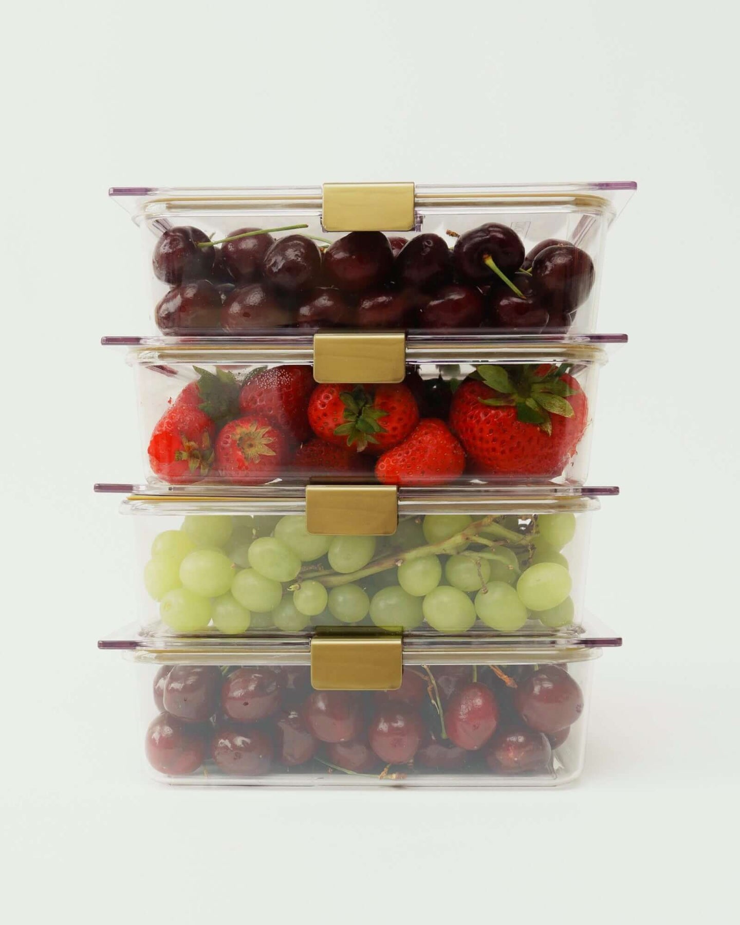 A studio shot showing four clear, rectangular food containers stacked vertically. The containers are filled with cherries, strawberries, green grapes, and dark red grapes. All containers have gold-colored locking clips.