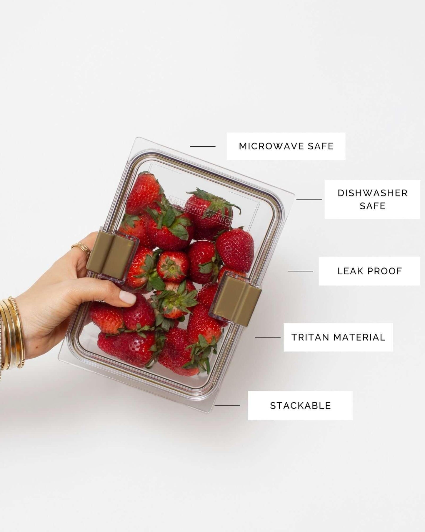 A hand wearing gold bracelets holds a clear, rectangular Tritan food container filled with fresh red strawberries. Feature labels highlight the container's attributes: Microwave Safe, Dishwasher Safe, Leak Proof, Tritan Material, and Stackable. The container has gold-colored locking clips on the sides.