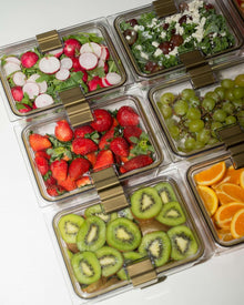 An overhead view of six clear, rectangular food containers with gold-colored clips, filled with colorful fresh food. Contents include sliced radishes on greens, salad with grapes and feta, strawberries, green grapes, sliced kiwi, and sliced oranges.