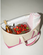 A large beige canvas cooler tote bag with a pink base and accents is shown open from above, revealing the white insulated interior. The bag is packed with picnic items, including a bottle of wine, a container of fresh strawberries, utensils, and a rolled-up pink and white blanket or towel.