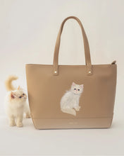A small, fluffy white Persian kitten sits next to a large cream tote bag that has a matching hand-painted portrait of the cat on the front.