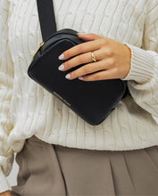 A close-up shot shows a person wearing a cream-colored cable-knit sweater and taupe pleated pants, holding a black leather crossbody or fanny pack. The bag has a small gold logo that reads "MODERN PICNIC" and a gold ring is visible on the hand holding the bag.