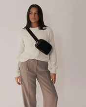 A young woman with dark hair, wearing a cream-colored cable-knit sweater and taupe trousers, models a black leather fanny pack/crossbody bag. The animation shows her adjusting the bag as she transitions from having it resting on her hip to opening the zipper to reveal the white insulated lining.