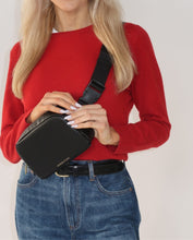 A blonde woman wearing a red sweater and blue jeans with a black belt is wearing a black leather fanny pack as a crossbody bag across her chest.