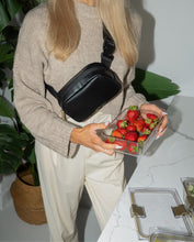A person with long blonde hair, wearing a tan knit sweater and light-colored trousers, stands in a kitchen. She is wearing a black fanny pack across her body and holding a clear food container full of fresh red strawberries over a white marble counter. A large leafy plant is visible to the left.