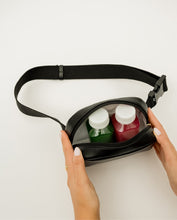 A pair of hands with a white manicure holds open a black fanny pack to reveal two small pressed juice bottles inside, one with a green liquid and one with a red liquid. The bag has a black strap and is against a white background.
