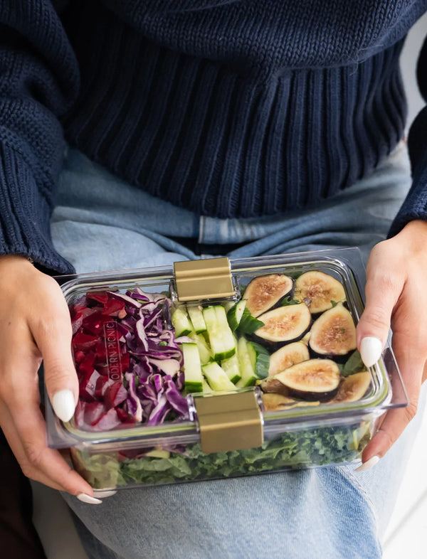 Person holding a clear glass meal prep container filled with a fresh salad with figs and cucumber.
