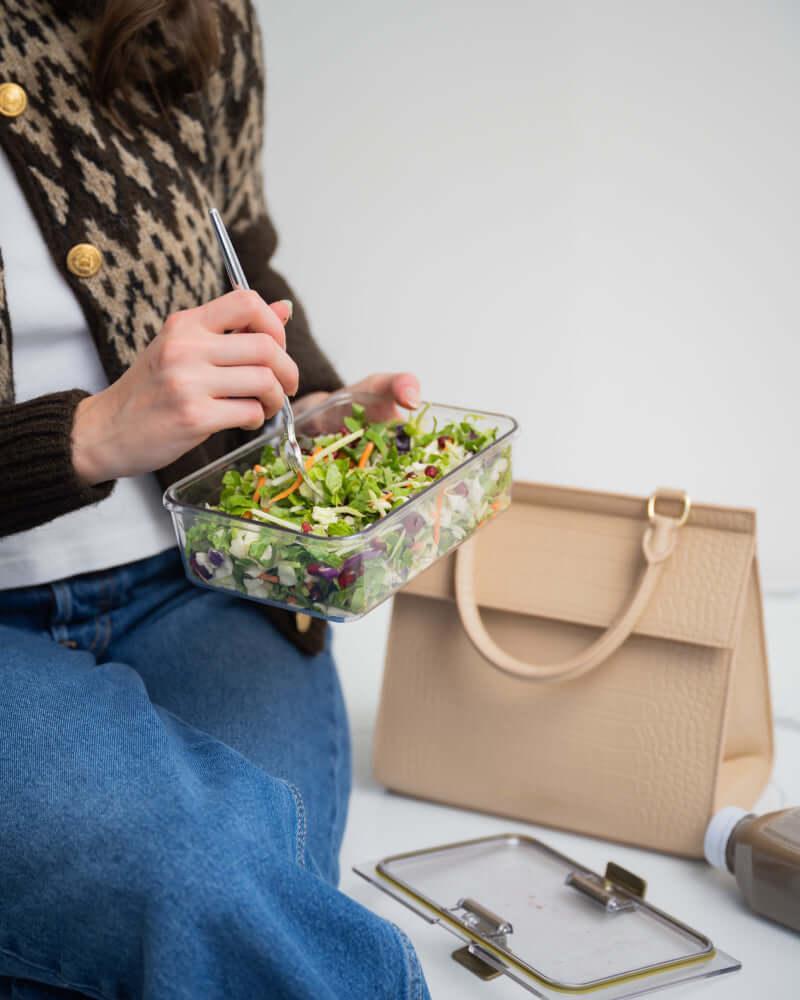A person wearing a brown patterned cardigan and blue jeans is sitting down, eating a salad from a clear food container with a fork. Next to her is a cream colored handbag with a crocodile-embossed texture, and the detached container lid rests on the floor.