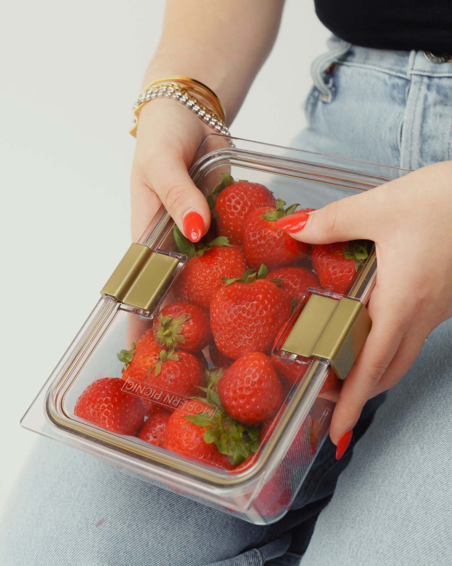 A close-up of a person's hands with bright red manicured nails and gold bracelets, holding a clear, rectangular food container filled with fresh red strawberries. The container has gold locking clips.