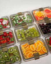 An overhead view of nine clear, rectangular food containers with gold-colored clips, all filled with a variety of fresh, colorful food. Contents include sliced kiwi, oranges, grapes, cherries, strawberries, grapefruit, and prepared salads.