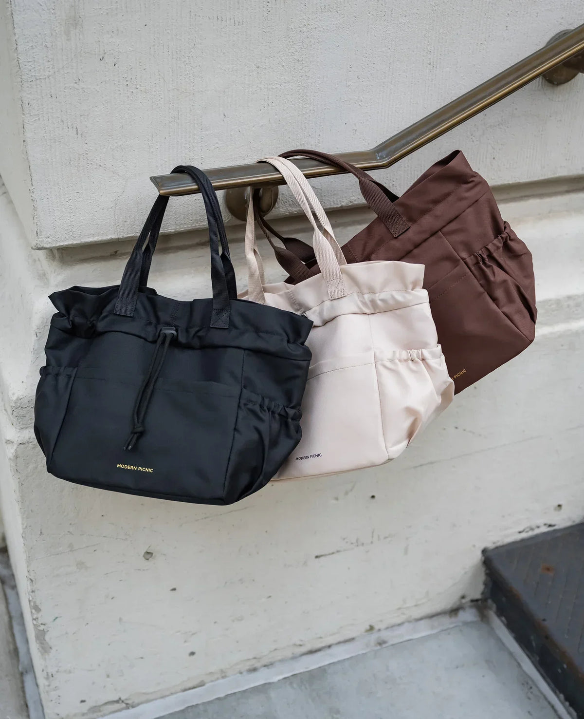 Three Modern Picnic insulated lunch totes in black, cream, and brown hanging on a handrail.