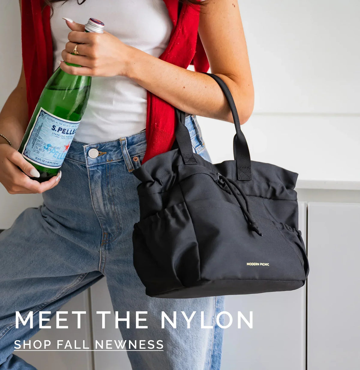 Woman in blue jeans and red cardigan holding a bottle of sparkling water and a black nylon tote bag.