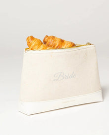 Cream canvas lunch bag with 'Bride' text, holding croissants, gold zipper, and vegan leather base