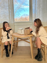 Two young girls sitting at a small table enjoying a snack, with a Stone Kids Mini and a Marigold Croc Kids Mini bag placed in front of them. Both girls are wearing cream outfits with black accents, and the scene is set in a bright room with a large window and rattan chairs, showcasing the lunch bags in a real-life, kid-friendly setting.