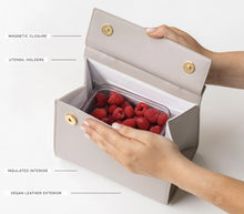 Open view of the Kids Mini Stone insulated bag by Modern Picnic, showing the stone-colored vegan leather exterior, insulated interior, magnetic closure, and utensil holders. A clear container filled with fresh raspberries is placed inside the bag, with hands holding the flap open on a white background.
