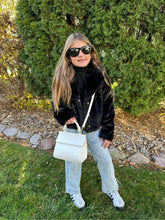Young girl standing outside on grass wearing sunglasses, a black sweater, jeans, and white sneakers, holding the Kids Mini Stone bag crossbody. Green hedges and rocks in the background create a casual outdoor setting, showcasing the Mini Stone bag styled on a kid.