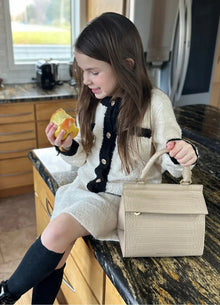 Young girl sitting on a kitchen counter eating an apple, wearing a cream and black outfit with knee-high socks, with the Kids Mini Stone bag placed beside her; real-life moment highlighting how the Mini Stone bag fits into everyday kid-friendly routines.