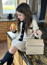Young girl sitting on a kitchen counter eating an apple, wearing a cream and black outfit with knee-high socks, with the Kids Mini Stone bag placed beside her; real-life moment highlighting how the Mini Stone bag fits into everyday kid-friendly routines.