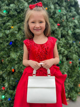 Smiling young girl dressed in a festive red dress with a matching red bow, standing in front of a decorated Christmas tree and holding the Kids Mini bag. A cheerful holiday scene showcasing the mini bag as a festive, kid-friendly accessory.