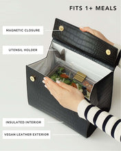 An overhead shot of a black handbag with a crocodile-embossed texture opened to display its function. A hand is reaching into the bag, which features a white insulated interior holding a clear container of salad. Labels point to the magnetic closure, utensil holder, insulated interior, and vegan leather exterior. The text overlay reads, "FITS 1+ MEALS."