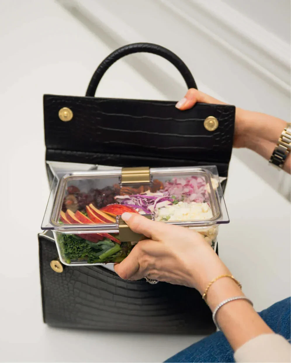 A close-up shot of a black crocodile-embossed handbag with the top flap open, showing a pair of hands placing a clear, partitioned container into the white insulated interior. The container is packed with colorful foods, including apples, red cabbage, and kale.