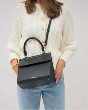A woman wearing a white cable-knit cardigan with gold buttons and blue jeans is carrying a structured black handbag with a crocodile-embossed texture. The bag is worn as a crossbody with its thin black strap and features a top handle and gold hardware.