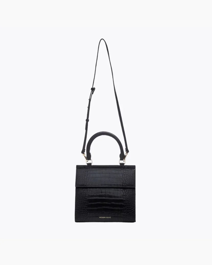 A front-facing studio shot of a structured black handbag made of crocodile-embossed vegan leather. The bag is suspended from its thin, adjustable black shoulder strap, with the top handle and gold hardware visible.