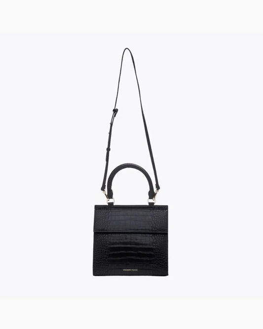 A front-facing studio shot of a structured black handbag made of crocodile-embossed vegan leather. The bag is suspended from its thin, adjustable black shoulder strap, with the top handle and gold hardware visible.
