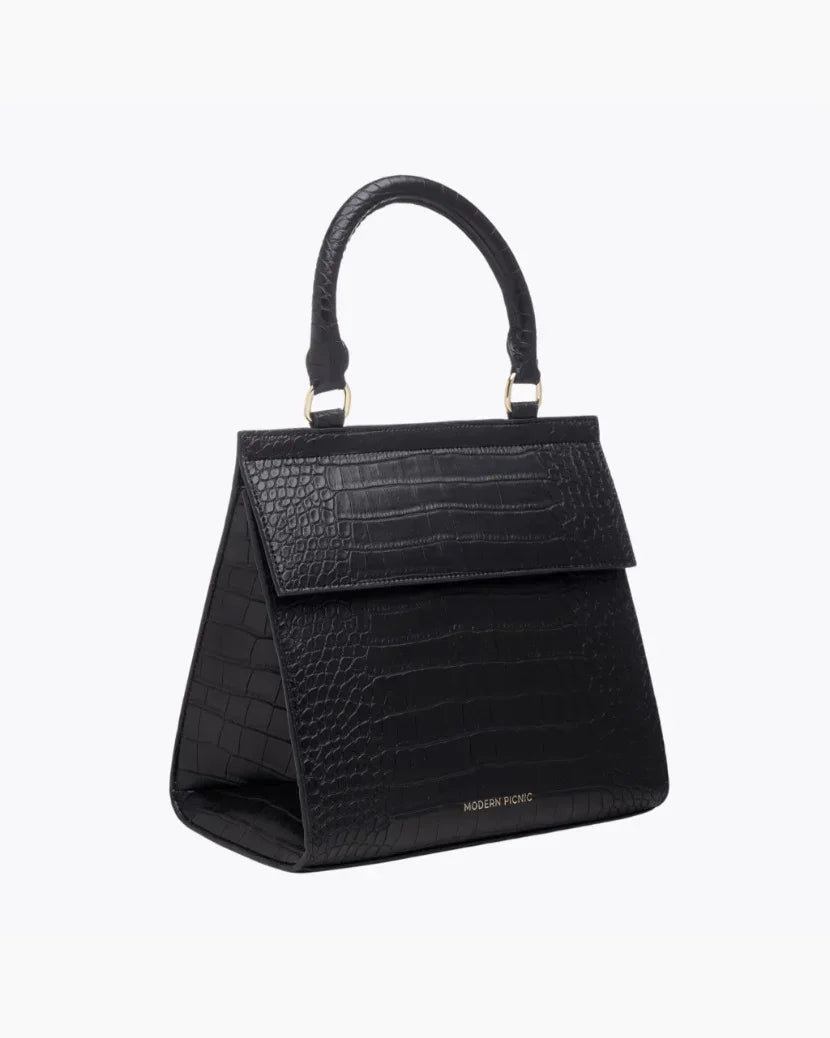 A three-quarter studio shot of a structured black top-handle handbag. The bag features an all-over crocodile-embossed texture, a prominent flap closure, and a visible gold "MODERN PICNIC" logo near the bottom front.