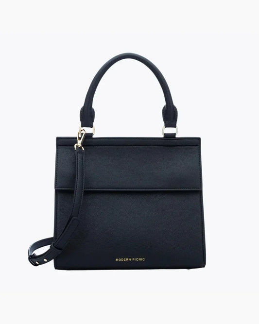 A front-facing studio shot of a structured black top-handle handbag. The bag is made of pebbled vegan leather and features a clean flap closure, a detachable matching shoulder strap, and gold hardware. The logo, "MODERN PICNIC," is subtly visible in gold near the bottom.