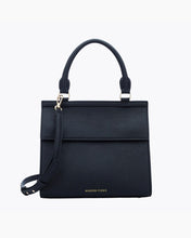 A front-facing studio shot of a structured black top-handle handbag. The bag is made of pebbled vegan leather and features a clean flap closure, a detachable matching shoulder strap, and gold hardware. The logo, "MODERN PICNIC," is subtly visible in gold near the bottom.