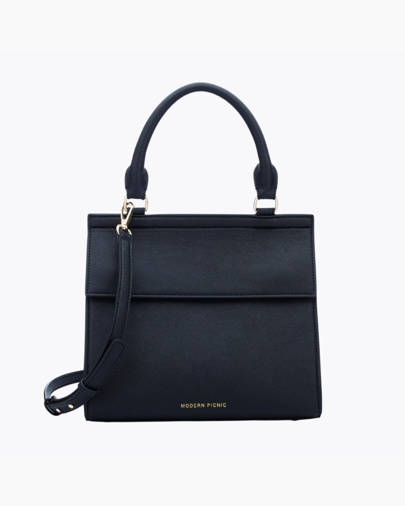 A front-facing studio shot of a structured black top-handle handbag. The bag is made of pebbled vegan leather and features a clean flap closure, a detachable matching shoulder strap, and gold hardware. The logo, 