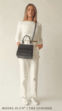 A full-body animation shows a blonde woman, wearing a white long-sleeve top and white trousers, modeling a structured black handbag made of pebbled vegan leather. The bag is worn both with the top handle and as a crossbody. The animation concludes with her opening the bag to reveal the white insulated interior holding a packed lunch container.