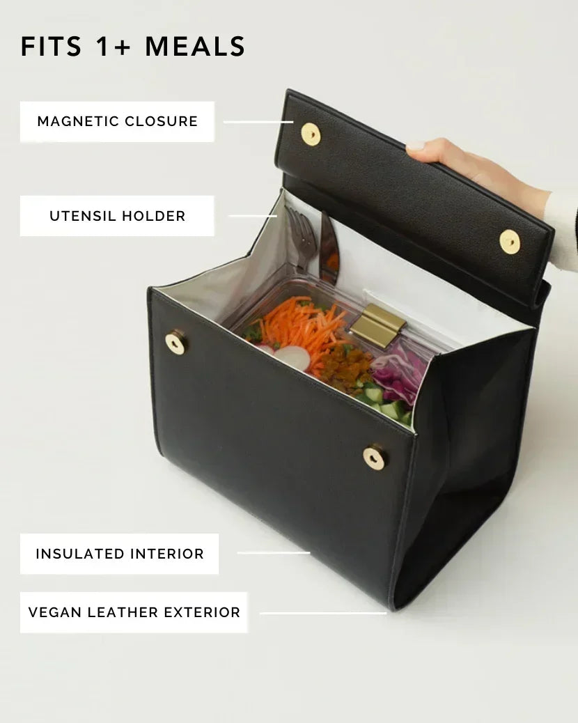 An overhead shot shows a black top-handle handbag made of pebbled vegan leather opened to display its function as an insulated lunch bag. Inside, the white insulated interior holds a clear container of salad with shredded carrots and purple cabbage, and a fork is visible in the utensil holder. Feature labels point out the magnetic closure, utensil holder, insulated interior, and vegan leather exterior. The text overlay at the top reads, "FITS 1+ MEALS."