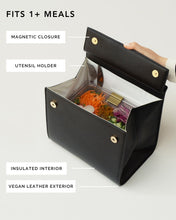 An overhead shot shows a black top-handle handbag made of pebbled vegan leather opened to display its function as an insulated lunch bag. Inside, the white insulated interior holds a clear container of salad with shredded carrots and purple cabbage, and a fork is visible in the utensil holder. Feature labels point out the magnetic closure, utensil holder, insulated interior, and vegan leather exterior. The text overlay at the top reads, "FITS 1+ MEALS."